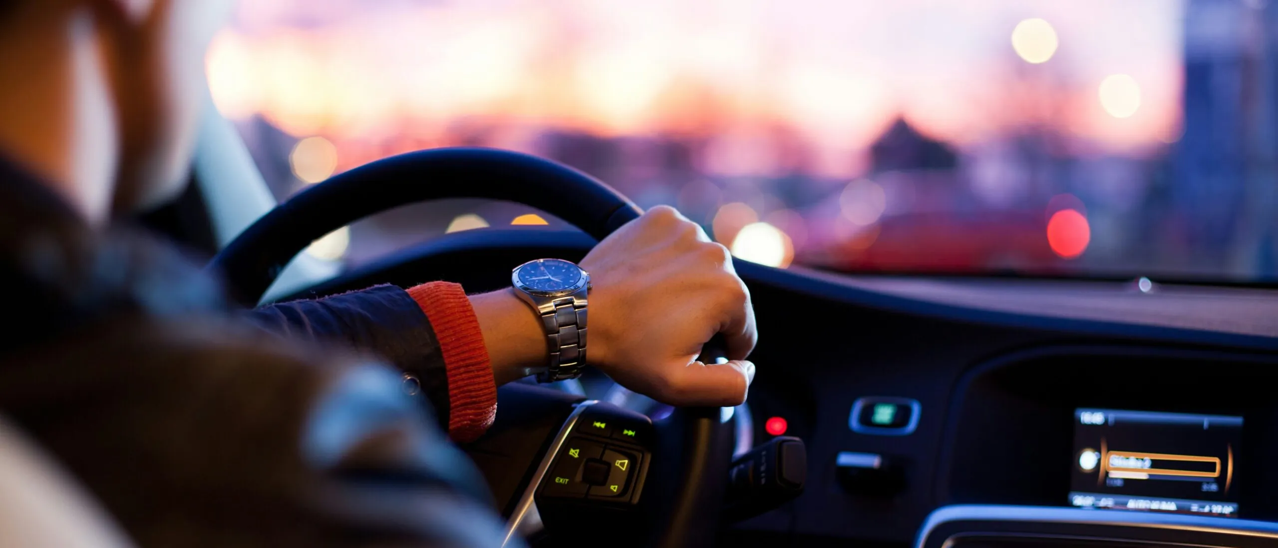 A driver at the wheel of a car at dusk, city lights ahead through the windscreen